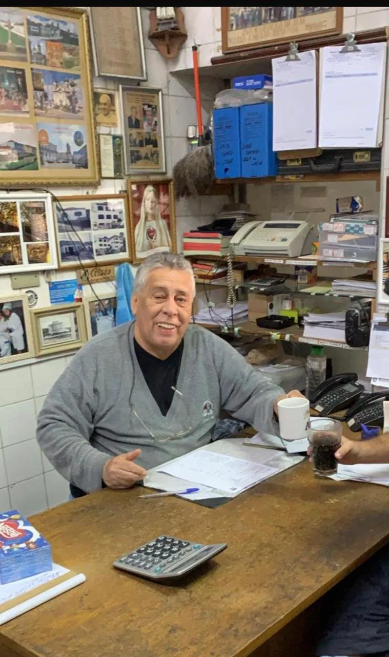 Celso Kersul, fundador da Vitrais Celso, sorridente à mesa do escritório da loja, cercado por fotos e lembranças na parede